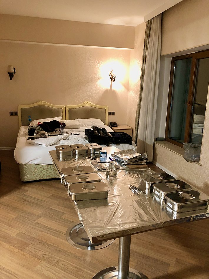 Das Bild zeigt ein Hotelzimmer mit einem großen Bett und einem Tisch, der mit mehreren Metallbehältern und etwas Zubehör bedeckt ist. Der Raum hat einen warmen Farbton und eine Fensterfront. Ein Koffer und Kleidung liegen auf dem Bett.
