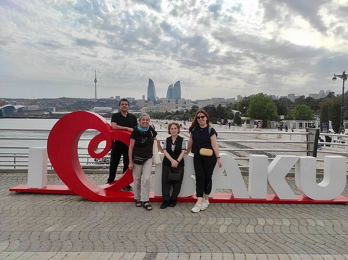 Vier Personen posieren vor einem großen roten Schriftzug "I ❤️ Baku" am Ufer. Die Stadtlandschaft mit modernen Gebäuden ist im Hintergrund sichtbar. Der Himmel ist teilweise bewölkt.
